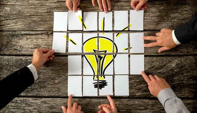 Manos juntan piezas que forman una bombilla amarilla sobre mesa de madera.