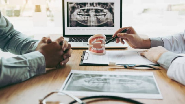 A dentist uses a pen to point to a dental model while reviewing dental X-rays during a patient consultation.
