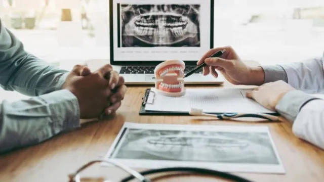 A dentist uses a pen to point to a dental model while reviewing dental X-rays during a patient consultation.