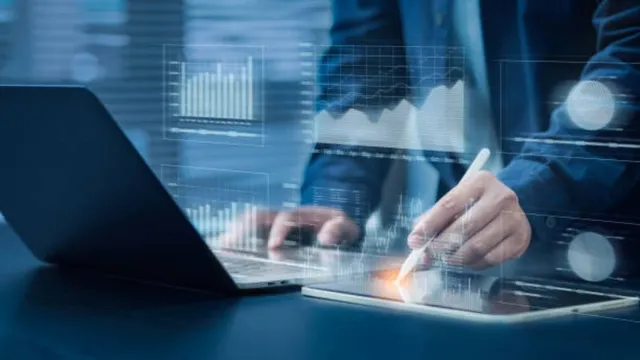 Close-up of a person using a laptop and a tablet with a stylus, featuring glowing digital overlays of business charts, graphs, and data analytics in a blue-toned office setting.