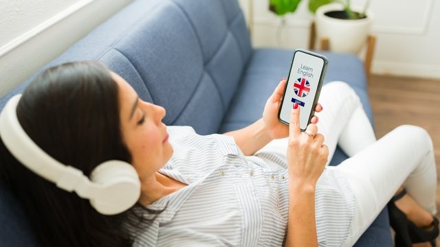 mujer tumbada en el sofá con auriculares y usando una app para aprender inglés en su móvil