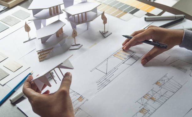 Hands work on an architectural project, holding a white paper model while sketching floor plans and elevations on a desk filled with color swatches and design tools.