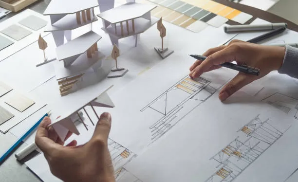 Hands work on an architectural project, holding a white paper model while sketching floor plans and elevations on a desk filled with color swatches and design tools.