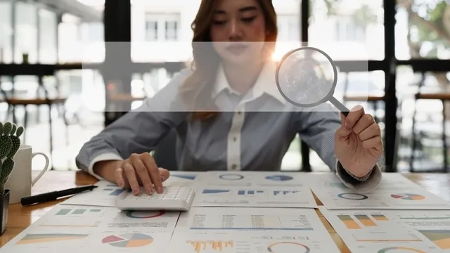 persona sentada frente a varios documentos con gráficos y estadísticas, sostiene una lupa
