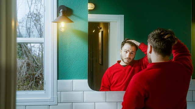 Joven con un suéter rojo mirándose en el espejo con una expresión preocupada, tocándose el cabello mientras contempla la pérdida de cabello