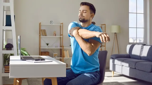 chico vestido con una camiseta azul y sentado en un escritorio frente a un ordenador portátil se estira durante un descanso laboral