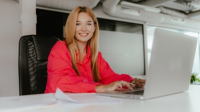 mujer sonríe a la cámara y tiene un portátil delante de ella sobre la mesa