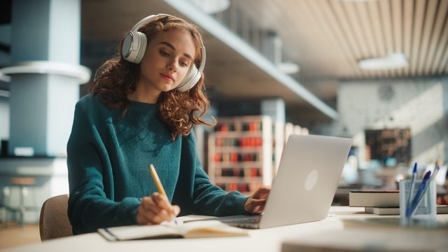 chica concentrada estudiando en la biblioteca con un ordenador portátil y los auriculares puestos