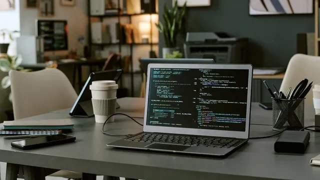 Ordenador portátil abierto que muestra el código de programación en la pantalla que se sienta en el escritorio en el espacio de trabajo moderno de la oficina con la taza de café, el smartphone, la tableta y los estantes en fondo, ninguna gente visible