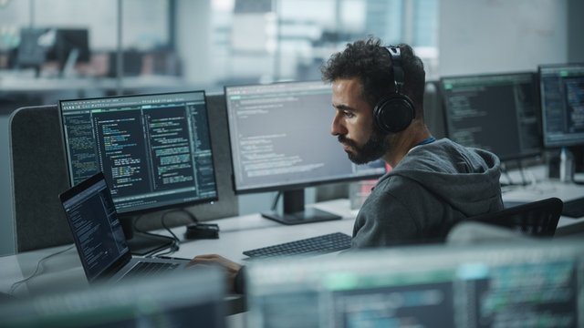 desarrollador de aplicaciones web con auriculares puestos, mientras trabaja con un ordenador de sobremesa y varias pantallas