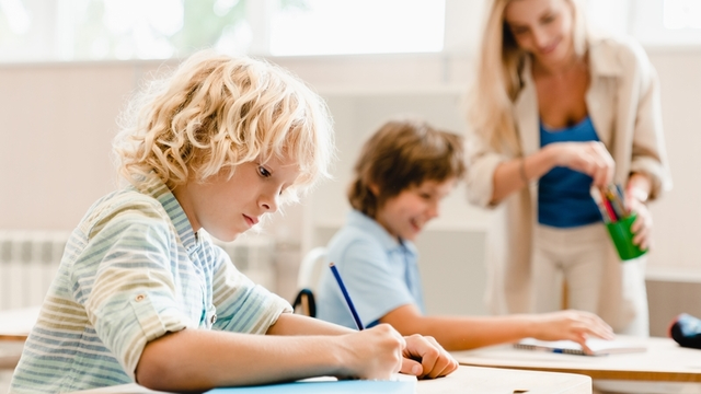 un niño concentrado escribiendo a lápiz en un cuaderno, mientras la profesora ayuda a otro compañero