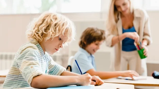 un niño concentrado escribiendo a lápiz en un cuaderno, mientras la profesora ayuda a otro compañero