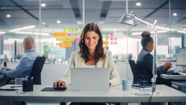 chica sonriente sentada frente a un ordenador portátil y trabajando en una oficina con dos personas más al fondo