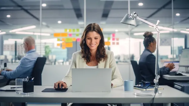 chica sonriente sentada frente a un ordenador portátil y trabajando en una oficina con dos personas más al fondo