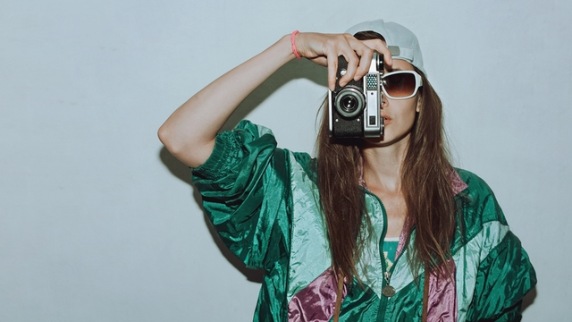 chica vestida con chaqueta vintage y gafas de sol retro sostiene una c&aacute;mara de fotos anal&oacute;gica