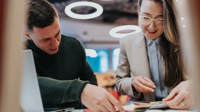 Dos compa&ntilde;eros de trabajo revisan documentos en un escritorio con una computadora port&aacute;til y papeles. Sonr&iacute;en y discuten ideas en un entorno de oficina moderno y brillante con luces de techo circulares.