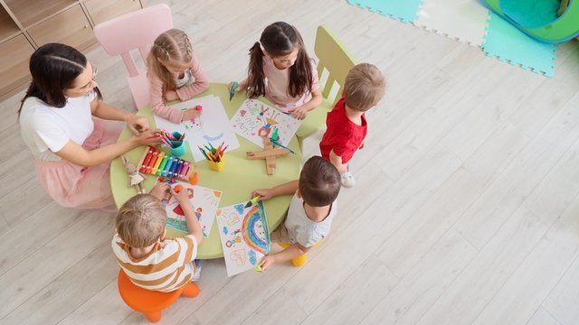 niños sentados alrededor de una mesa acompañados de una técnica superior en Educación Infantil y dibujando