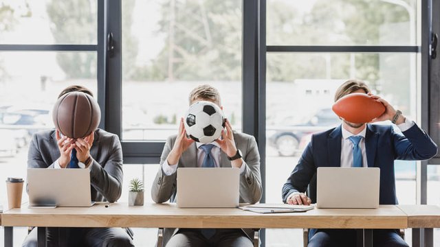 personas vestidas de traje sentadas frente a unos ordenadores portátiles y tapándose la cara con balones de diferentes deportes