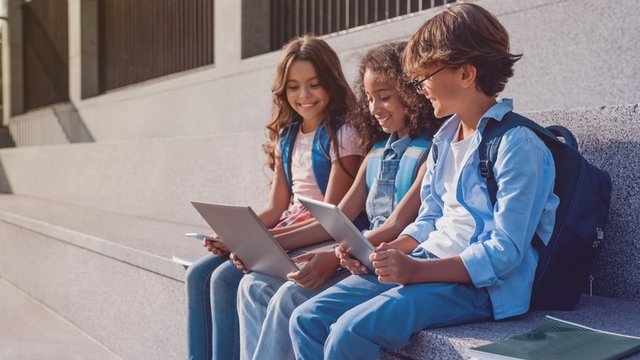 alumnos de primaria sentados en las escaleras y usando dispositivos digitales al aire libre