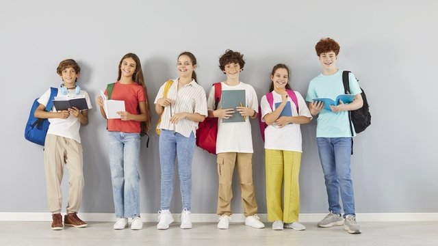 adolescentes sonrientes de pie delante de una pared gris sostienen material escolar