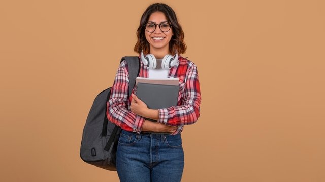 chica sonriente vestida con camisa a cuadros roja y blanca sostiene una pila de libros y lleva una mochila en un hombro