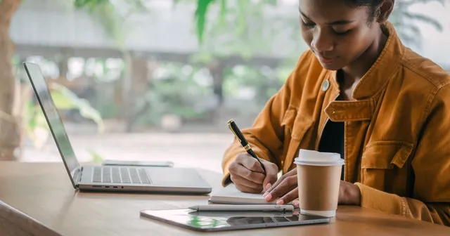 A young woman is writing in a notebook with a laptop and a cup of coffee on the table