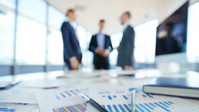 mesa cubierta de papeles con gráficos mientras 3 personas mantienen una conversación