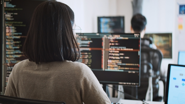 mujer sentada de espaldas trabaja en un proyecto de programación con varias pantallas de ordenador
