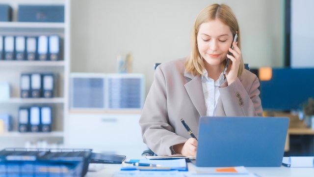 chica joven vestida de traje hace una llamada por teléfono, mientras apunta algo en un documento que tiene sobre el escritorio