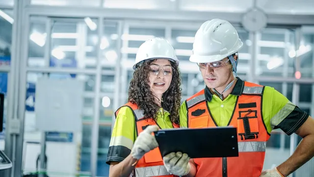 Dos ingenieros usan una tableta para programar un sistema de IA. Están en una fábrica inteligente futurista, revisando datos para robots industriales o fabricación de microchips.