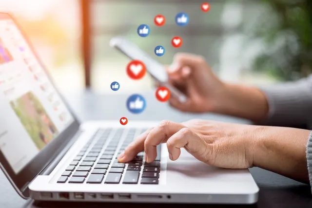 Persona gestionando redes sociales desde un ordenador portátil y un móvil con iconos de likes y corazones flotando en pantalla