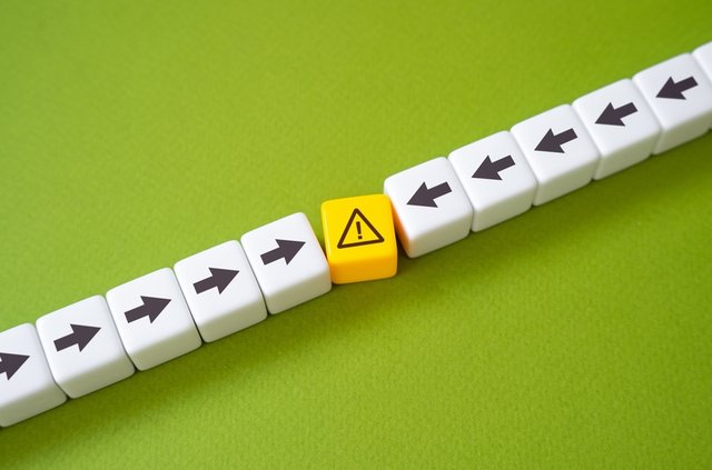 Cubos con flechas en direcciones opuestas chocan contra un símbolo de precaución en el centro, representando un conflicto de intereses.