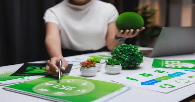 A person reviewing green ESG documents and sustainability diagrams on a desk with a small moss ball and a laptop.