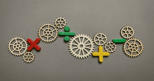 Wooden cogs interconnected with plastic math signs on a gray surface.