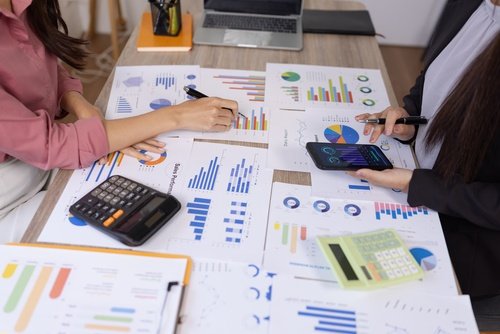 Two colleagues analyzing printed business charts and graphs with pens and a calculator on a desk.