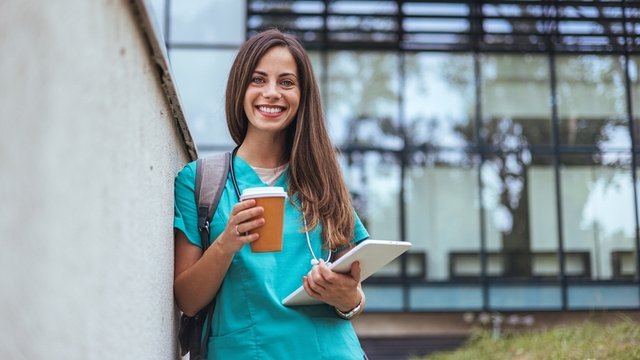 estudiante de enfermería sonriendo y sosteniendo un vaso de café enfrente de la facultad