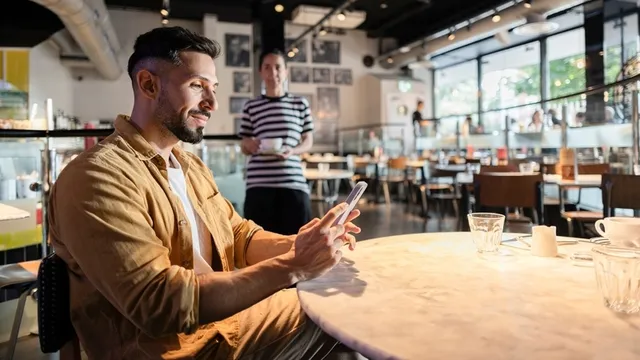 Hombre joven que usa el smartphone en el restaurante de Londres, Cliente sonriente que disfruta de la tecnología móvil en un café elegante, con un camarero que sirve en el fondo