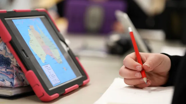 mano de un estudiante sostiene un lápiz, toma notas en un papel y observa un mapa en una tableta