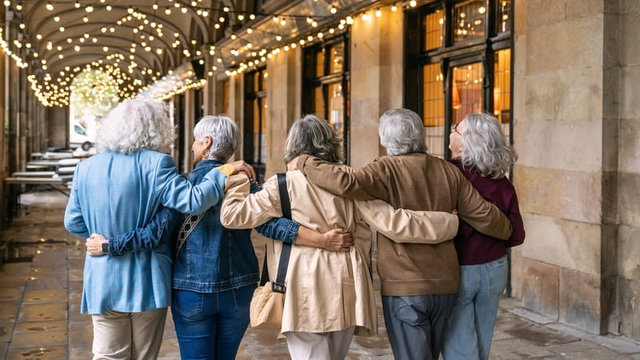 Un grupo diverso de personas mayores, caminando brazo a brazo a lo largo de una arcada iluminada, compartiendo un momento de estilo de vida retirado